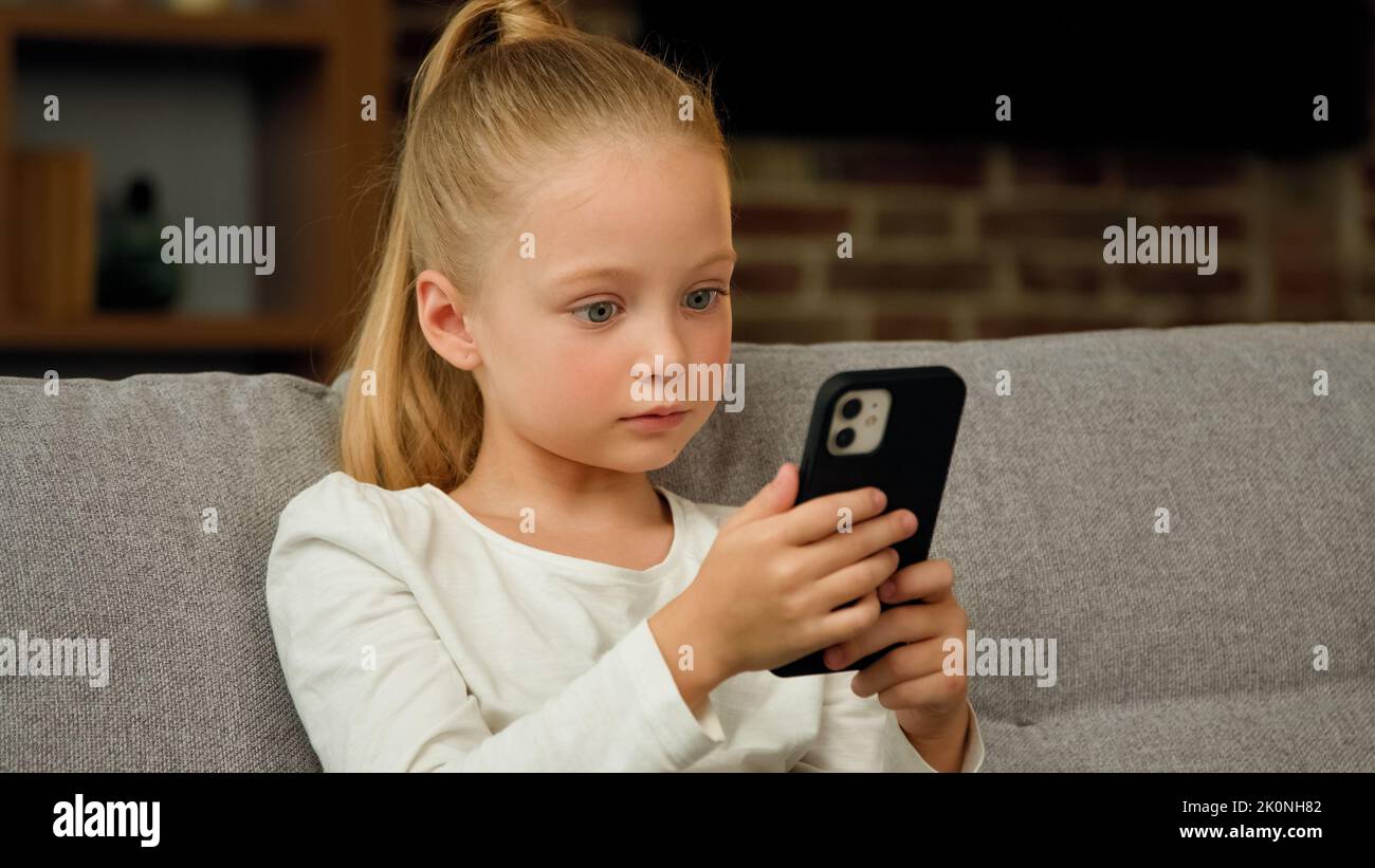 Beautiful pretty small girl pupil caucasian kid child daughter blogger sit on sofa at home use smartphone play mobile games chatting with classmates Stock Photo