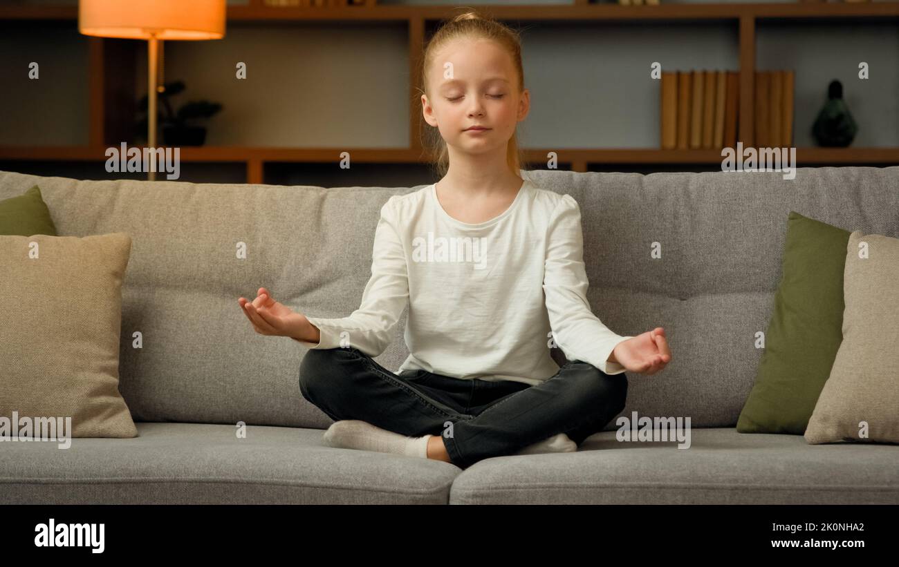 Little cute caucasian girl young healthy child blonde kid preschooler meditate sit on sofa at home calm mindful peaceful daughter do yoga breathing Stock Photo