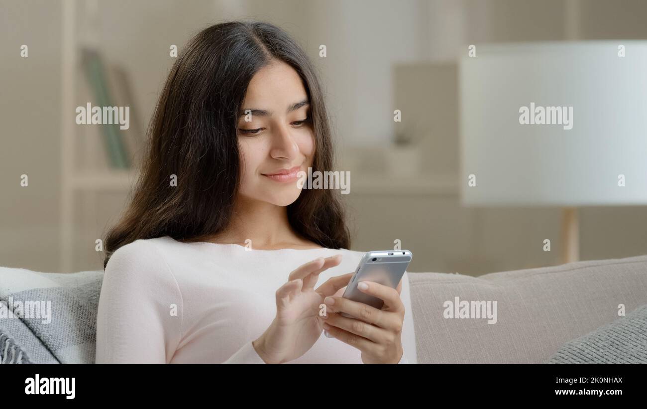 Female young user freelancer calm attractive girl arab hispanic woman sits on sofa at home scrolls inform pages on telephone device chatting makes Stock Photo