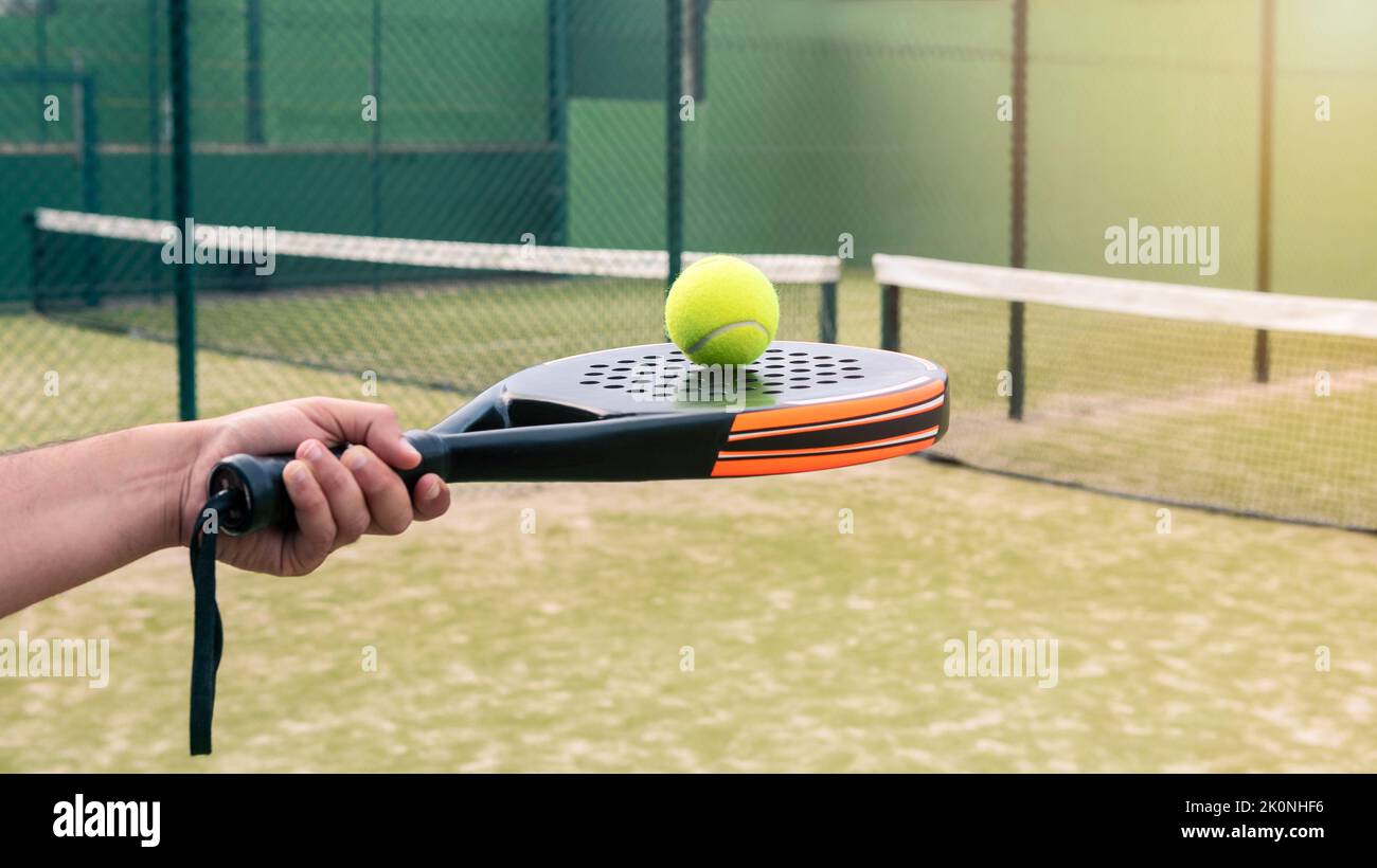 Professional monitor of padel holding a black racket with a yellow ball on top. Class to student on outdoor tennis court. Man paddel player playing a Stock Photo