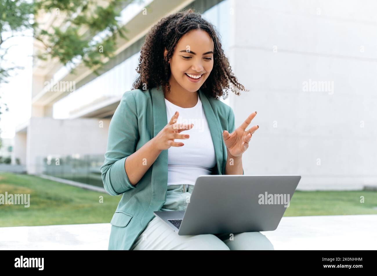 Friendly mixed race young business woman, sitting outdoors with a laptop near the business center, talking with clients or coworkers by video call, smiles. Online video conversation outdoors Stock Photo