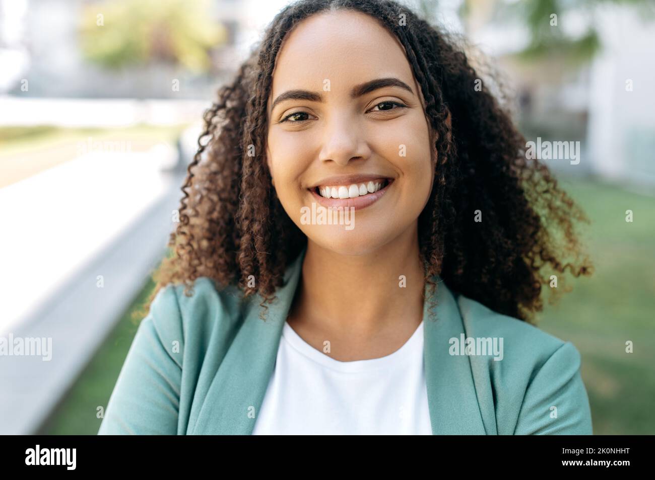 Close-up photo of beautiful positive confident successful hispanic or brazilian young woman with curly hair, business lady, in stylish elegant clothes, standing outdoors, looking at the camera, smiles Stock Photo
