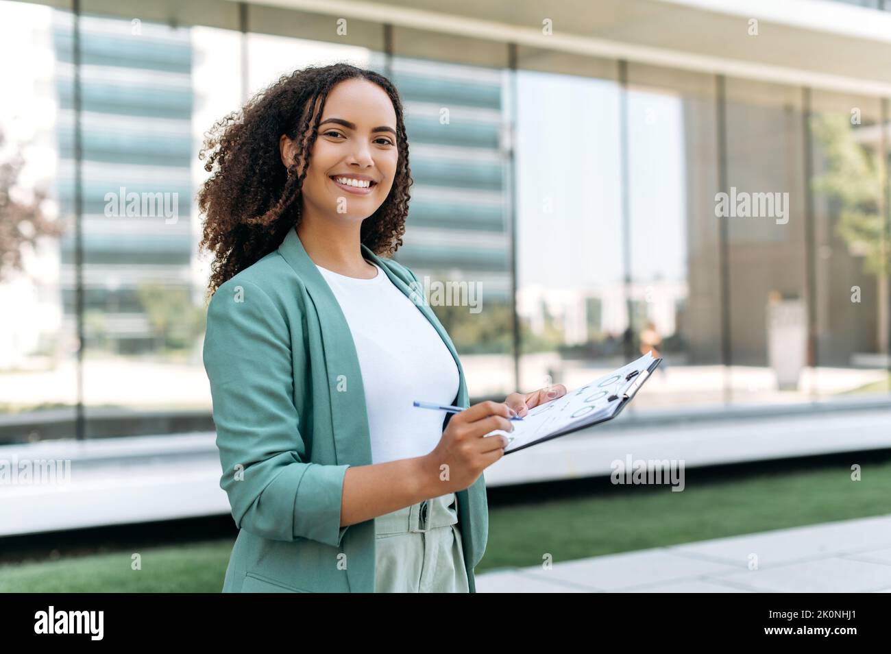 Portrait of lovely positive confident successful mixed race young woman with curly hair, business lady, trading broker, in elegant wear, standing outdoors with financial charts, looks at camera, smile Stock Photo