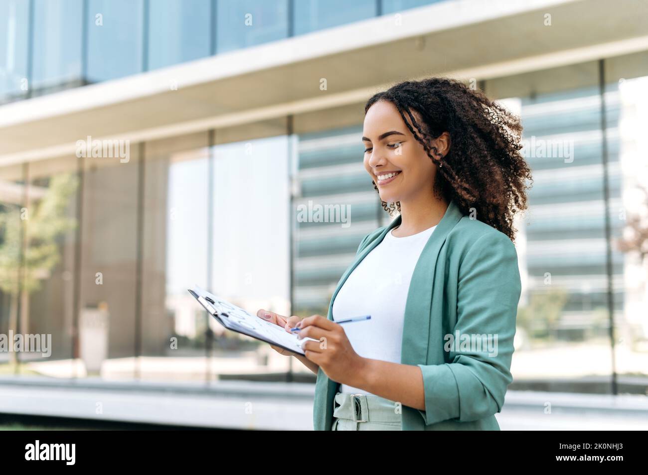 Beautiful confident successful latino or brazilian young woman with curly hair, business lady, financial expert, in elegant wear, standing outdoors with financial charts, analyze graphs, smiling Stock Photo