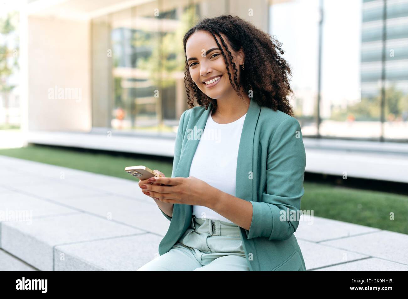 Lovely young brazilian or latino business woman, wearing elegant suit, holding cell phone sitting outdoors, using gadget for online messaging, checking email, browsing websites, look at camera, smiles Stock Photo
