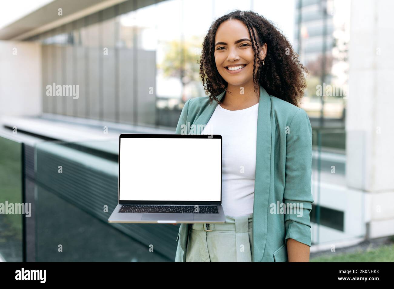 Happy successful pretty mixed race woman with curly hair, stylishly dressed, stands outdoors, holds an open laptop with blank white mock-up screen in hand, looks at camera, smiles friendly Stock Photo