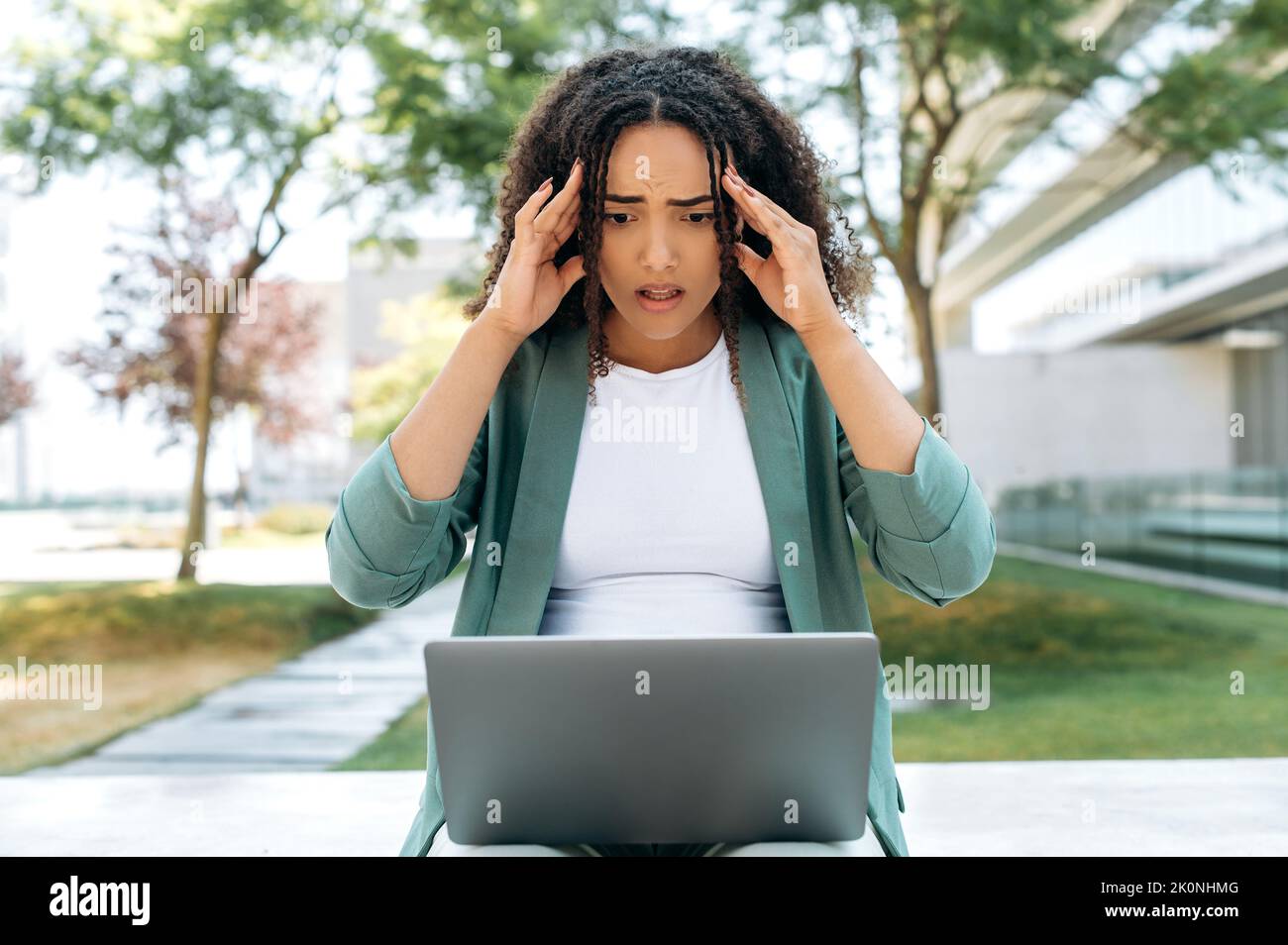 Frustrated mixed race young business woman, elegantly dressed realtor, manager, broker, sit outdoors with laptop, looking at screen in confusion, received unexpected news, holds her hands on her head Stock Photo