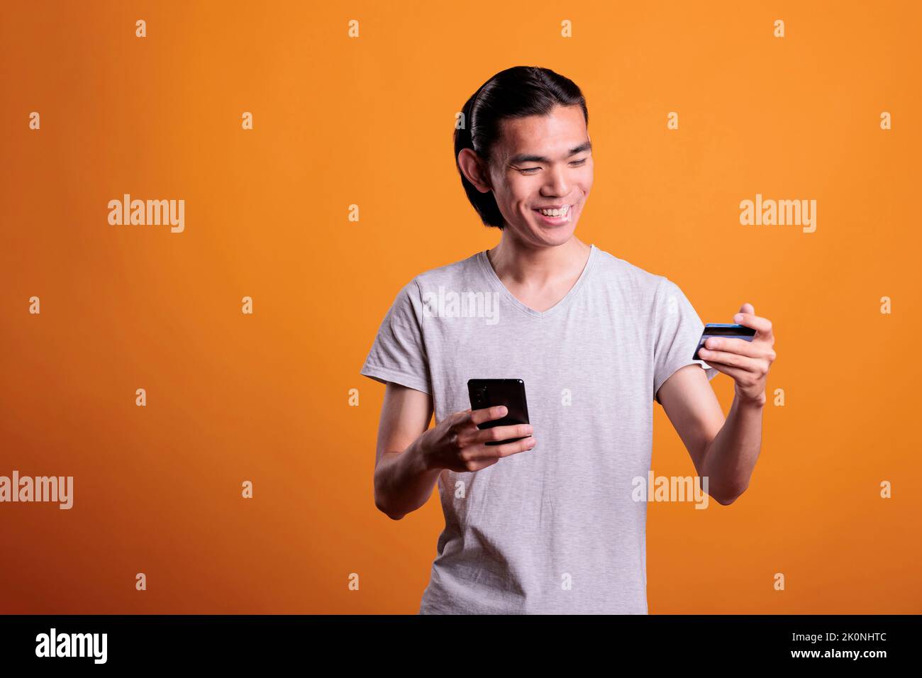 Smiling young man buying products in internet shop, making order using smartphone app. Asian teenager holding debit card and searching goods in online store, e commerce concept Stock Photo
