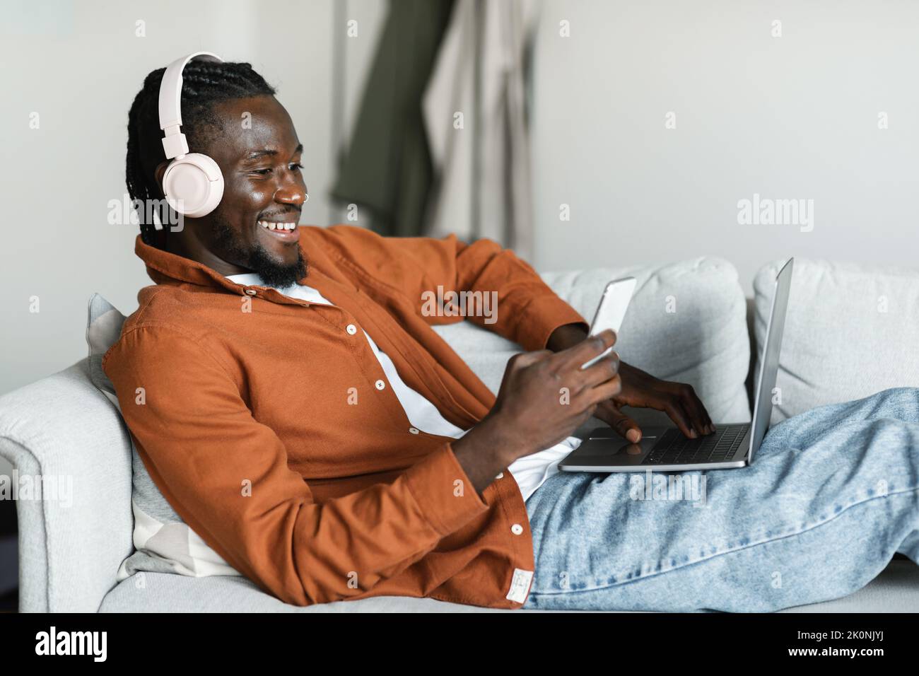 Smiling black man lying on couch with pc, using cellphone and wearing headset, taking break from remote work Stock Photo