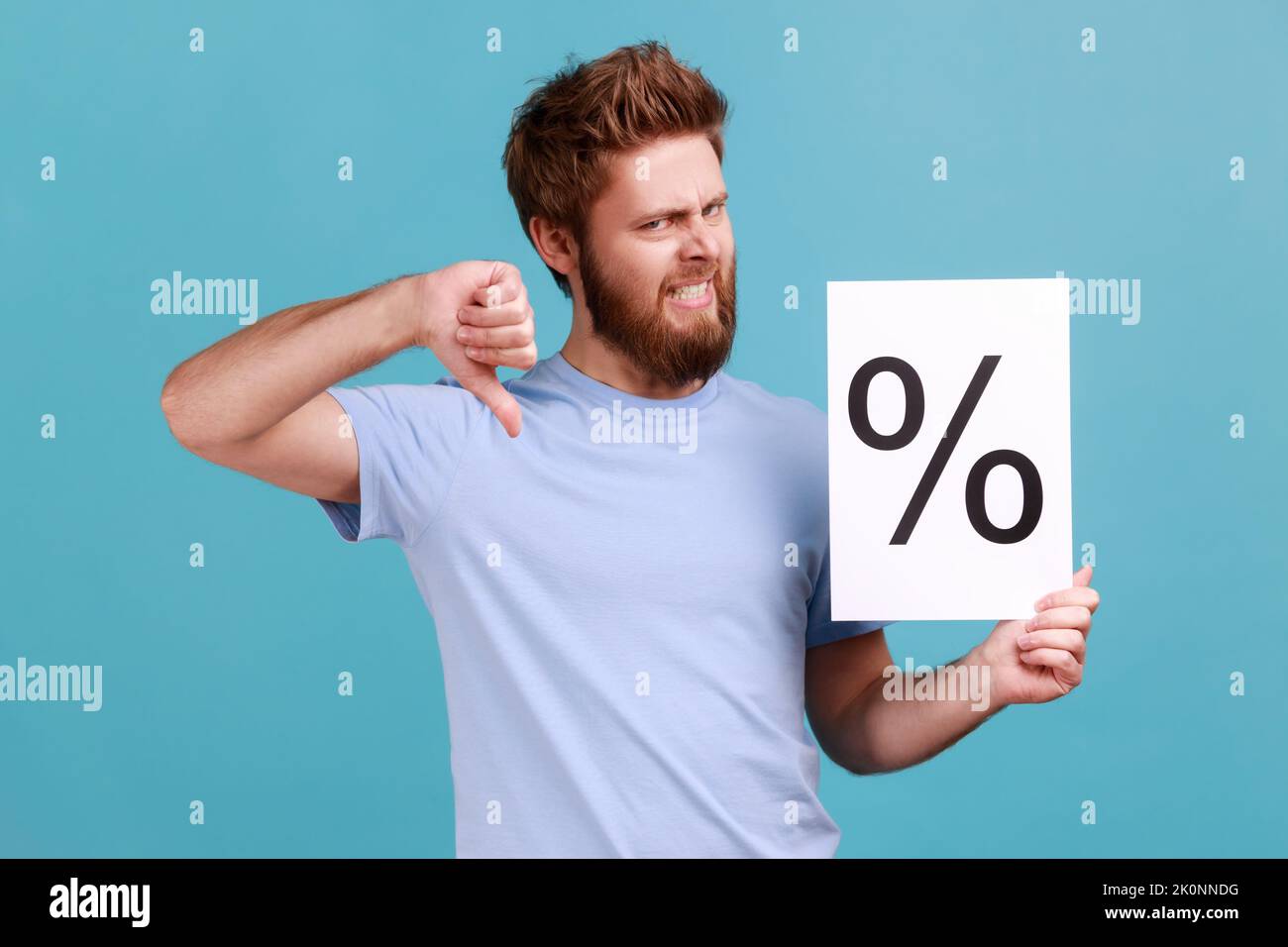 Portrait of handsome young adult bearded man holding paper with percent sign, looking at camera with frowning face and showing thumb down. Indoor studio shot isolated on blue background. Stock Photo