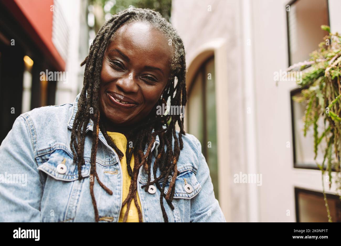 Ethnic woman with dreadlocks smiling happily while standing outdoors. Cheerful senior woman going out in casual clothing. Stock Photo