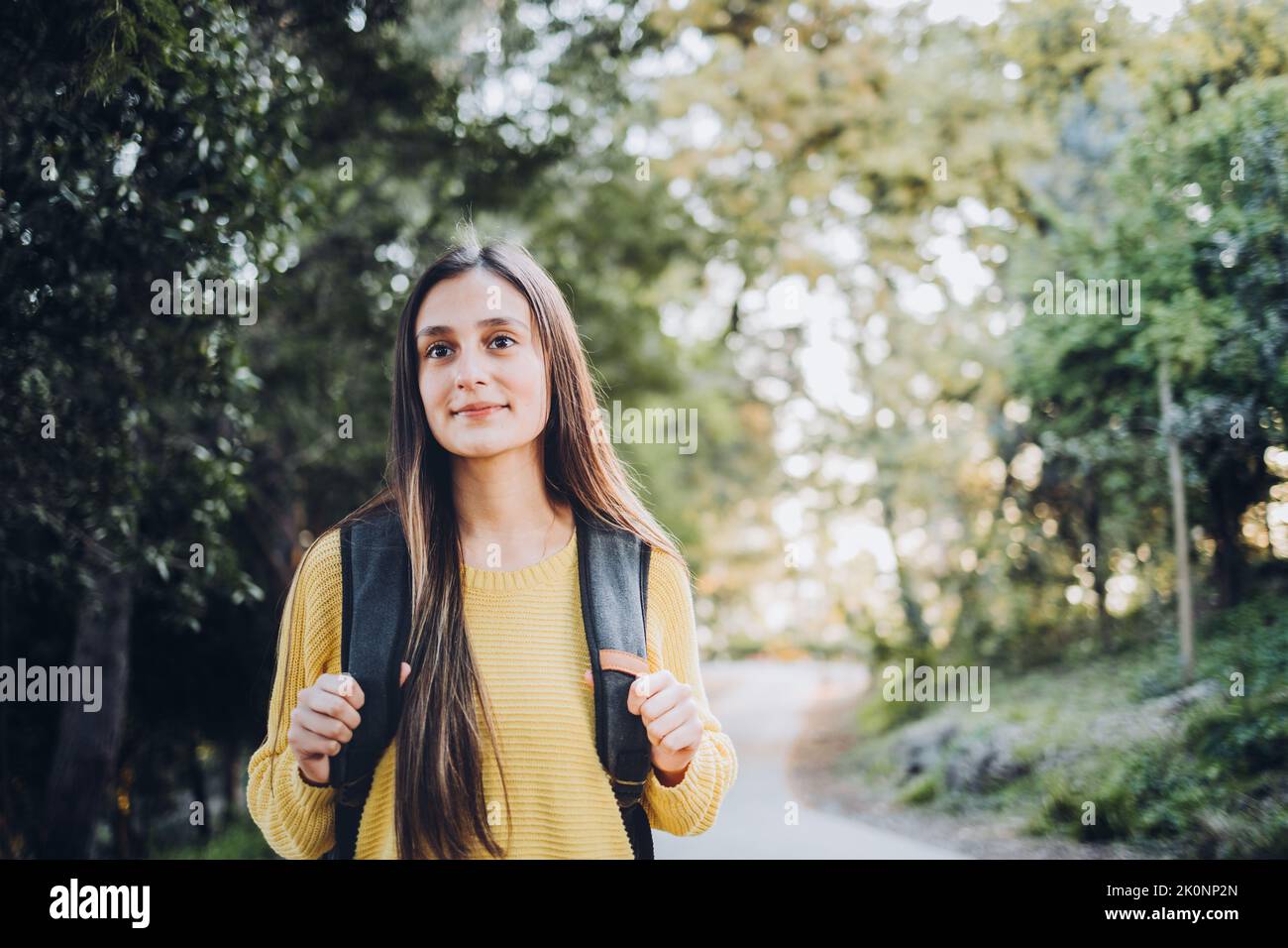 Smiling female university student wearing a yellow sweater and a backpack in the campus park road. Stock Photo