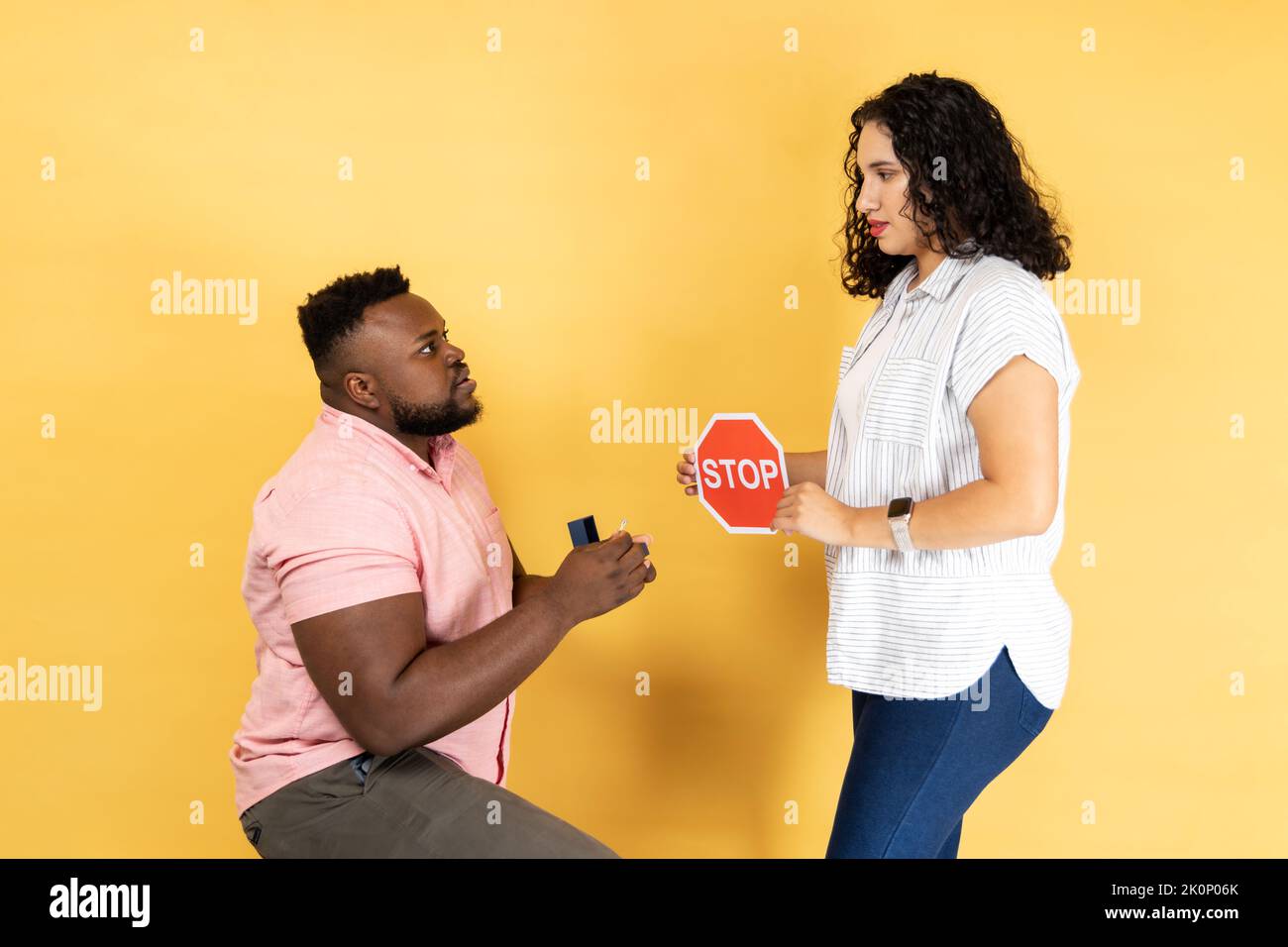 Portrait of young couple in casual clothing standing together, man making proposal to his girlfriend, woman showing red stop sign to him, refusing. Indoor studio shot isolated on yellow background. Stock Photo