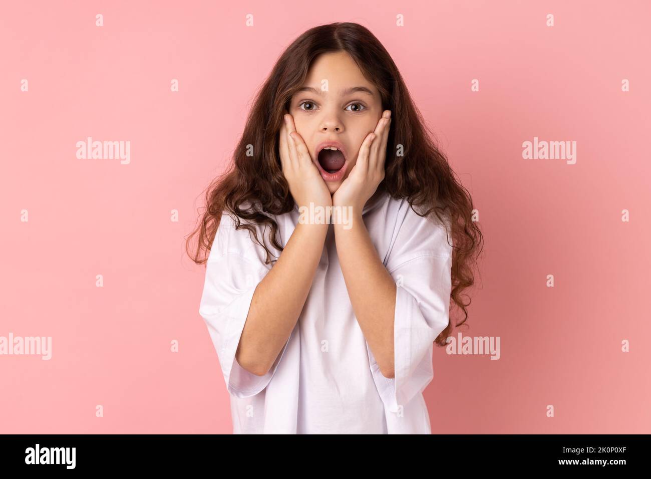 Portrait of amazed little girl wearing white T-shirt standing with hands on cheeks, looking at camera with big eye, saying wow. Indoor studio shot isolated on pink background. Stock Photo