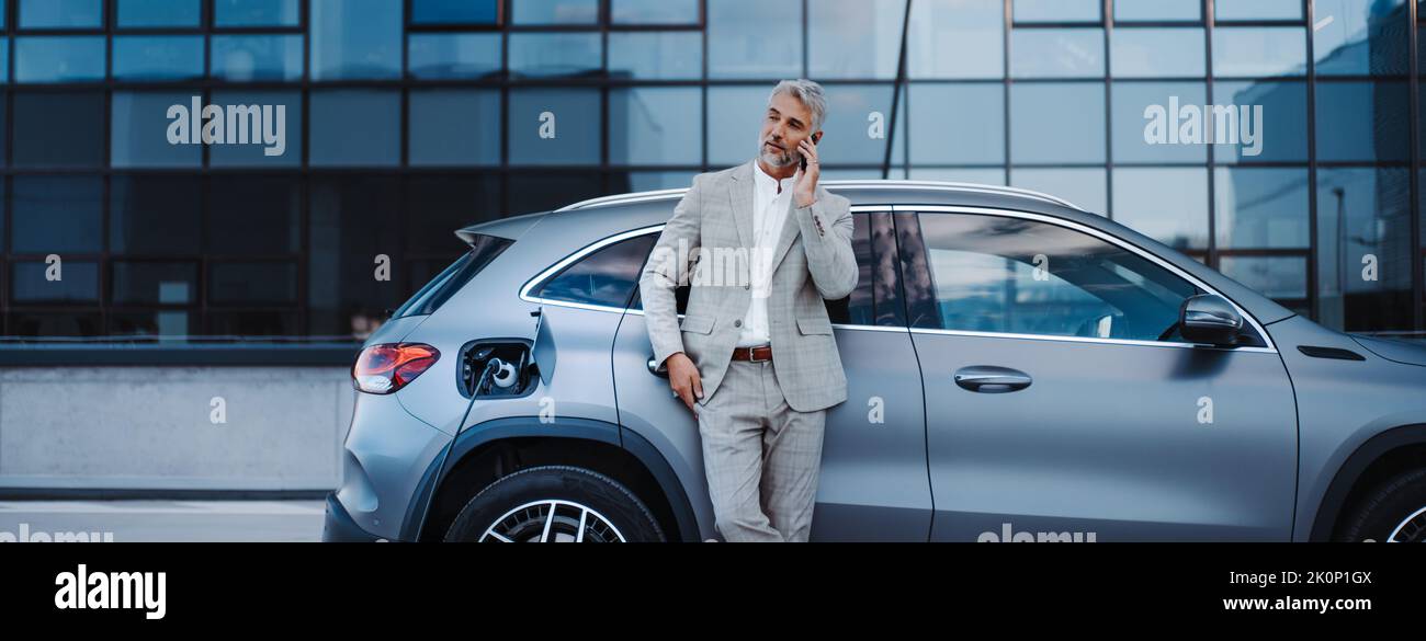 Businessman holding smartphone while charging car at electric vehicle charging station, closeup. Stock Photo