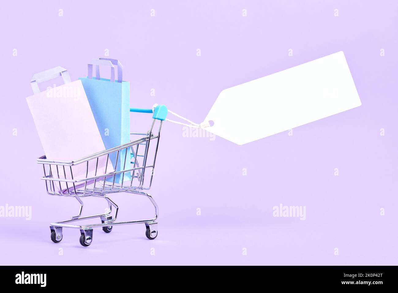 Shopping cart full of bags with a blank label on a pastel lilac background. Minimalist design with copy space. Concepts: market deals, seasonal sales Stock Photo