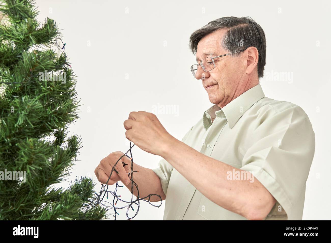 Hispanic senior man concentrated while decorating a Christmas tree, putting a string of led lights on it. Stock Photo