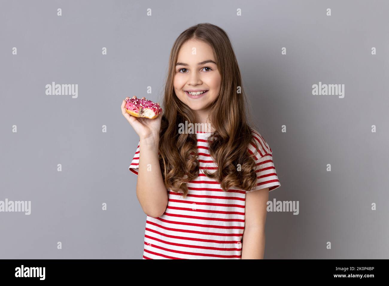 Portrait of delighted happy adorable little girl wearing striped T-shirt delicious sugary donut and smiling genuinely to camera. Indoor studio shot isolated on gray background. Stock Photo
