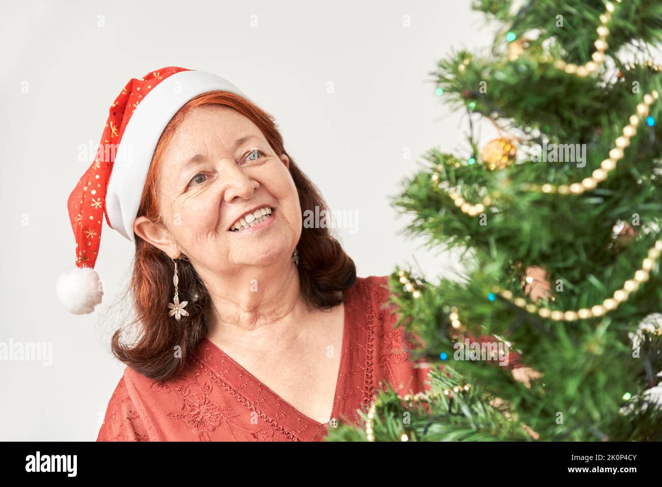 Happy senior hispanic woman smiling while decorating a Christmas tree wearing a red Santa Claus hat. The joy of the holidays. Stock Photo