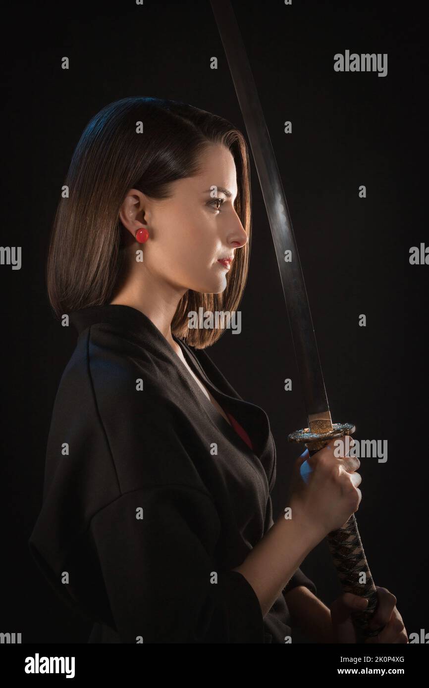 Portrait in profile of a young woman who holds a katana in her hands in front of her on a black background Stock Photo