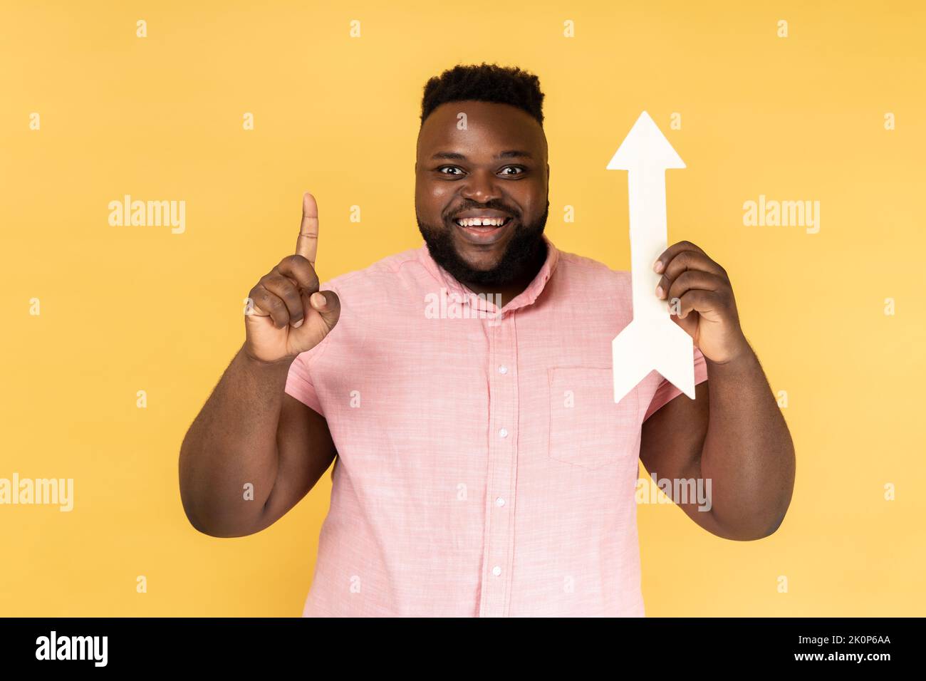 Portrait of satisfied delighted happy man wearing pink shirt holding arrow pointing up, raising finger, having idea, growth and increase concept. Indoor studio shot isolated on yellow background. Stock Photo