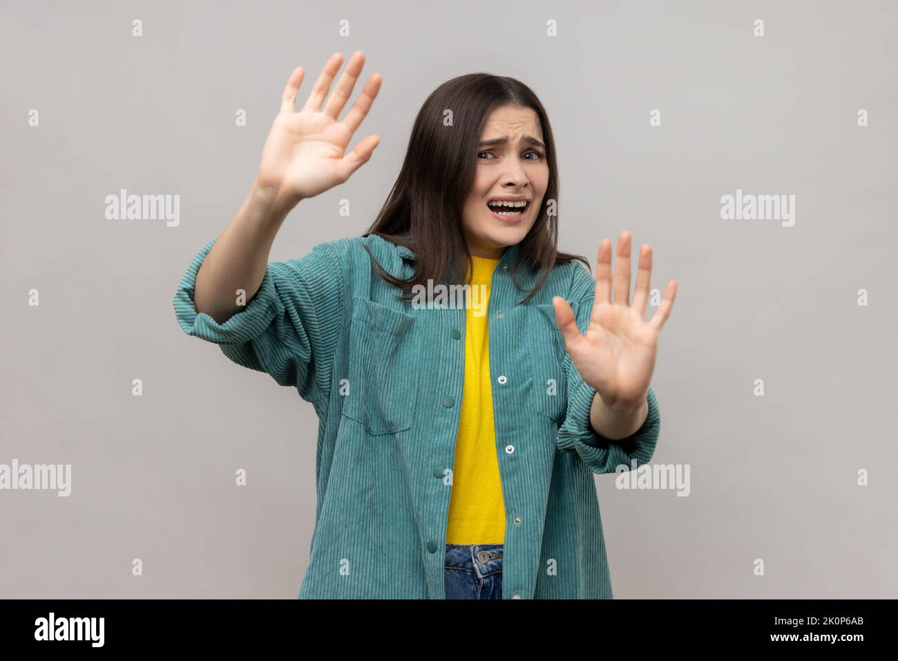 Portrait of scared young adult dark haired woman gesturing stop with palms and looking surprised with frightened eyes, wearing casual style jacket. Indoor studio shot isolated on gray background. Stock Photo
