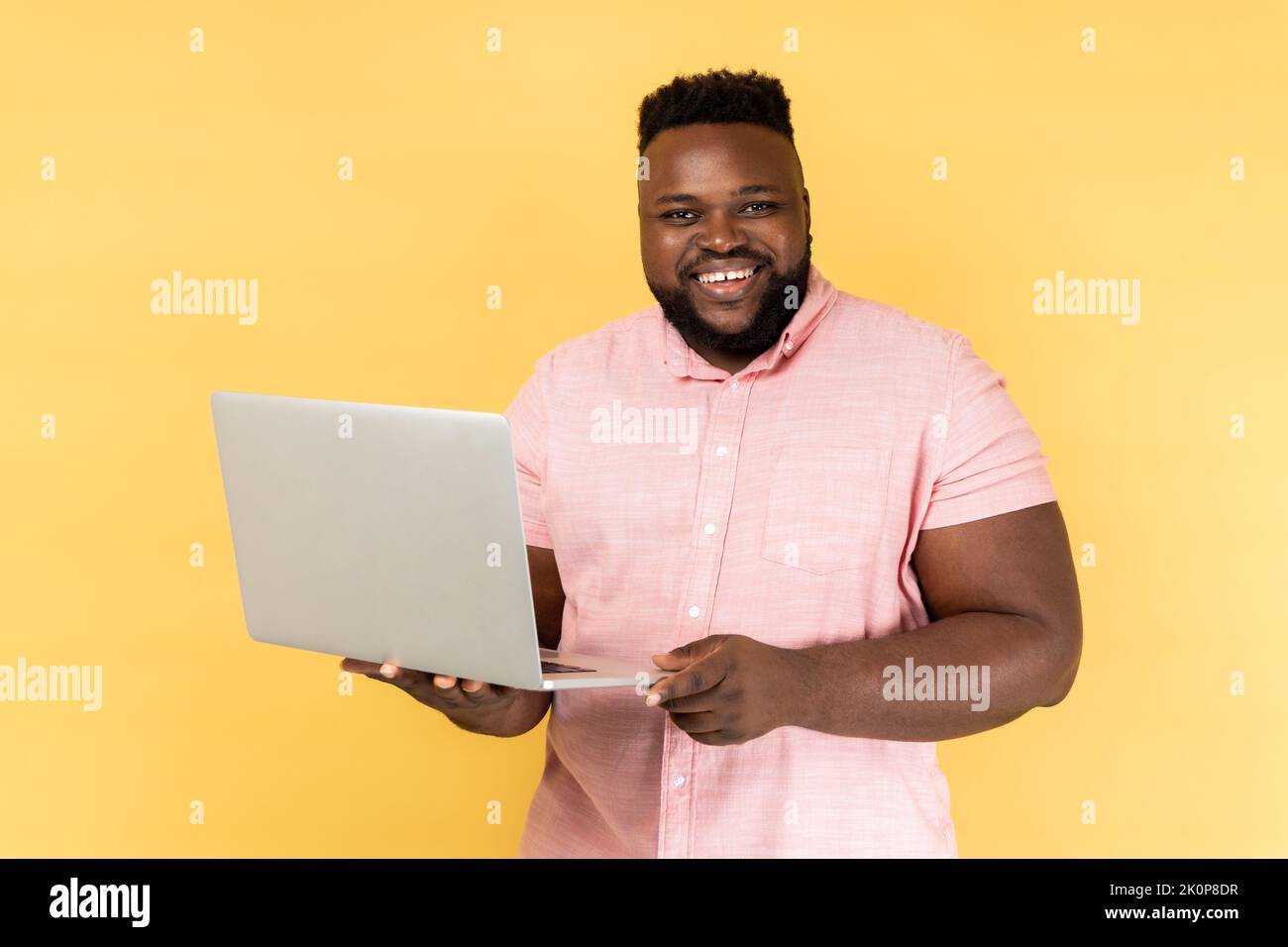 Portrait of smiling freelancer man wearing pink shirt standing and holding laptop, satisfied with teleworking, likes his job, looking at camera. Indoor studio shot isolated on yellow background. Stock Photo