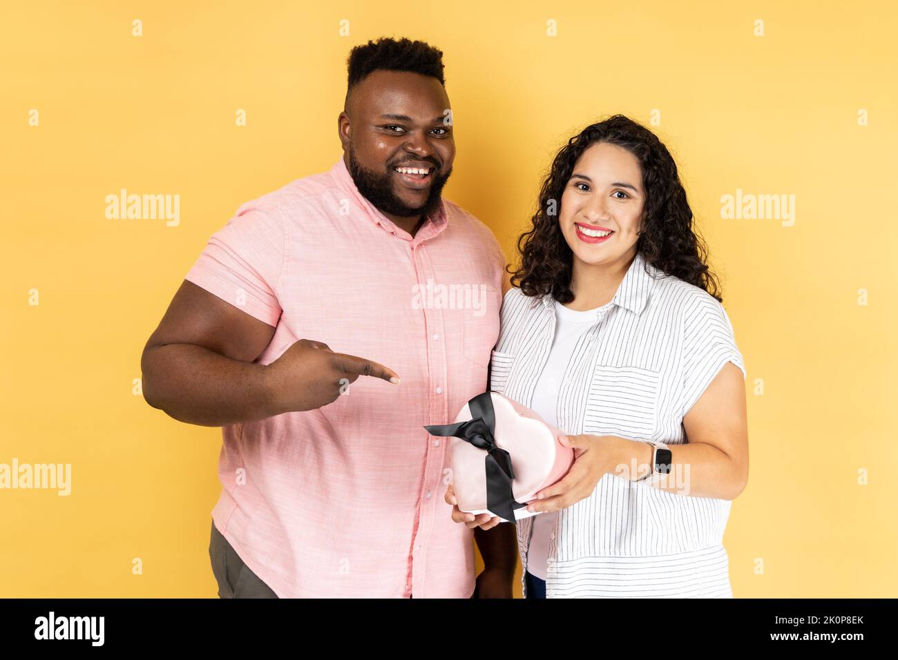 Portrait of delighted smiling young couple in casual clothing standing together, man pointing at present box in her wife hands, celebrating holiday. Indoor studio shot isolated on yellow background. Stock Photo