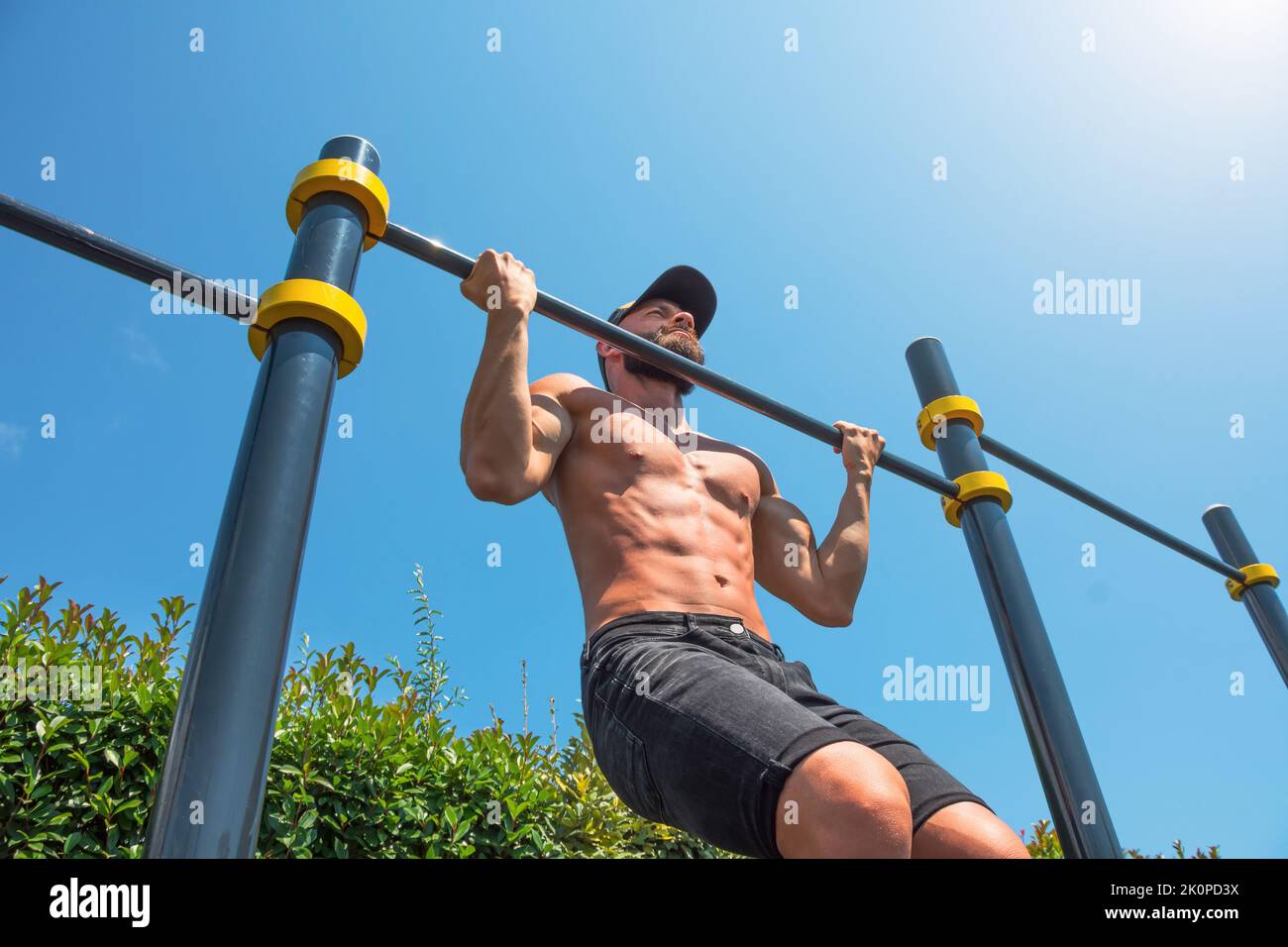 Muscular man in a cap doing reverse pull-up on the horizontal bar in the park outdoors Stock Photo