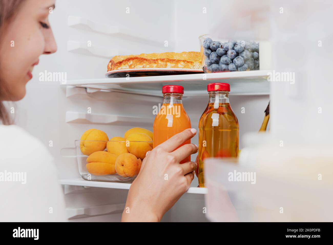 Woman hand taking, grabbing or picks up juice glass bottle out of open refrigerator shelf or fridge drawer full of fruits, vegetables, banana, peaches, yogurt. Healthy food diet, lifestyle concept Stock Photo