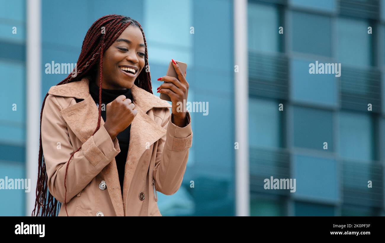 Excited enthusiastic surprised young woman winner standing outdoors reading good news on phone celebrating victory rejoicing in success winning Stock Photo