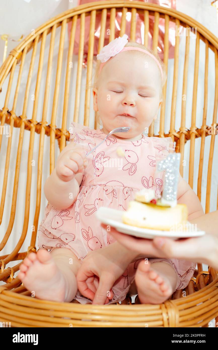 Vertical barefoot infant close eyes in pink dress and flower on head sit on chair and tasting birthday cake. One year Stock Photo