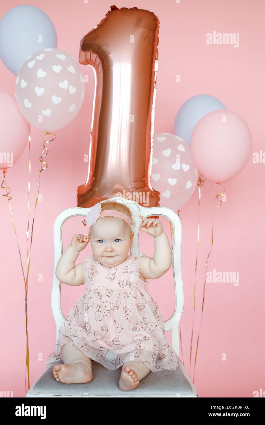 Vertical little joyful fortunate barefoot one year baby in pink dress and flower on head sitting on chair near balloons Stock Photo