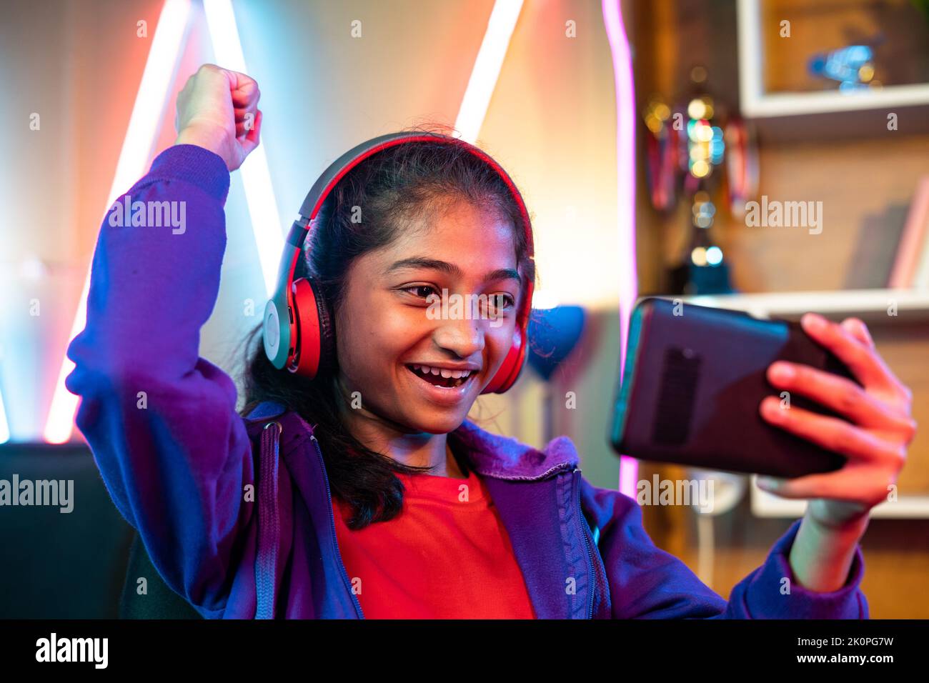 Excited teenager girl with headset celebrating win while playing video game on mobile phone at home - concept of achievement, competition and Stock Photo