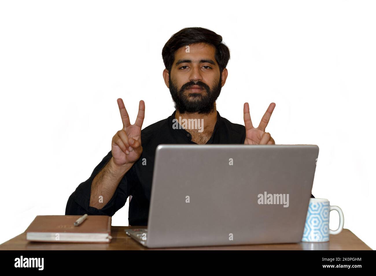 A bearded young man sitting on a chair and showing victory sign with both hands. Laptop, diary and coffee mug on table. Selective Focus on eye. Stock Photo
