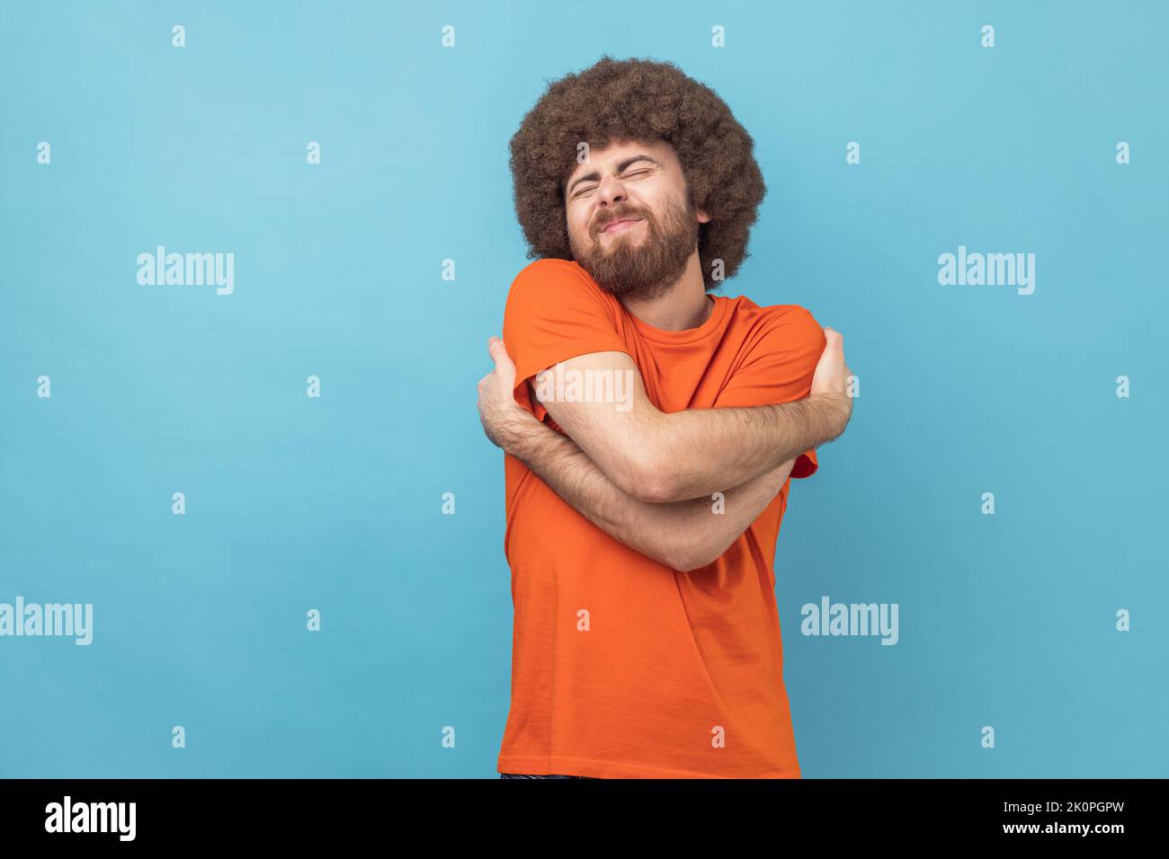 Portrait of selfish narcissistic man with Afro hairstyle embracing himself and smiling with expression of great ego, pleasure and self-esteem. Indoor studio shot isolated on blue background. Stock Photo
