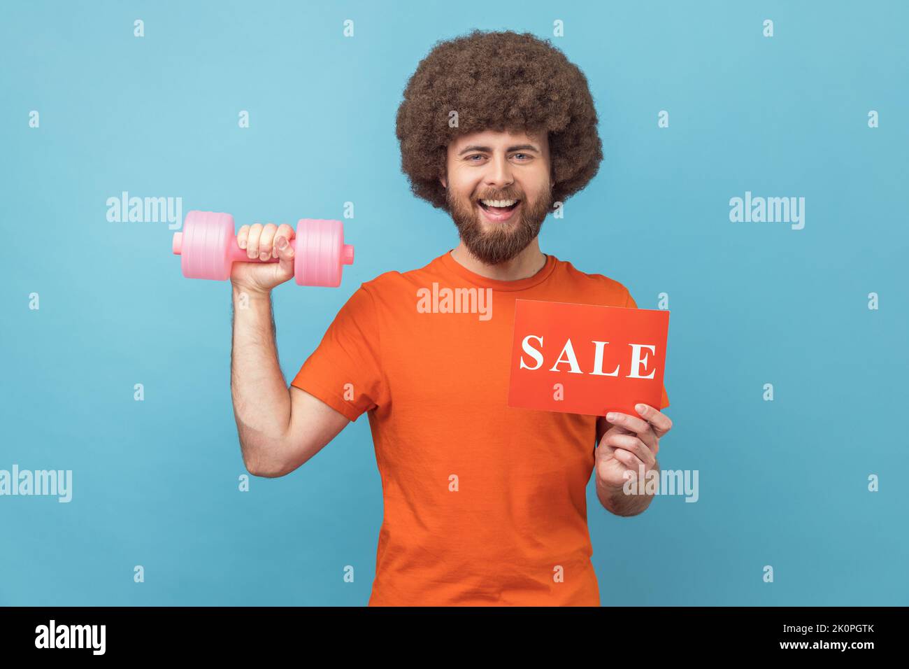 Portrait of satisfied man with Afro hairstyle wearing orange T-shirt holding dumbbell and card with sale inscription, sales for fitness workout. Indoor studio shot isolated on blue background. Stock Photo