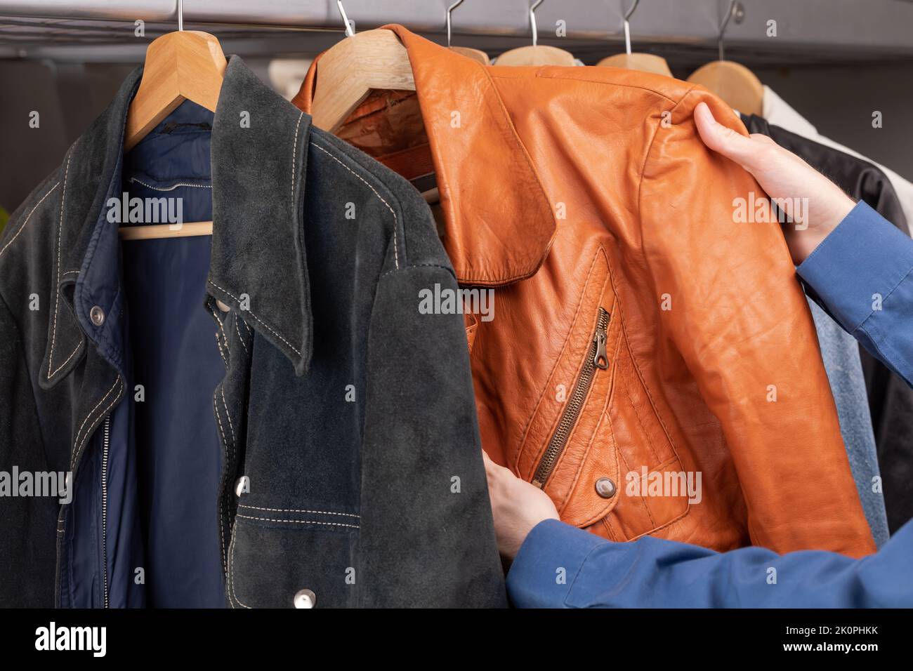 Man checks orange leather jacket in a second hand store. Various vintage jackets hang on clothing rack. Thrifting and sustainability in clothing conce Stock Photo