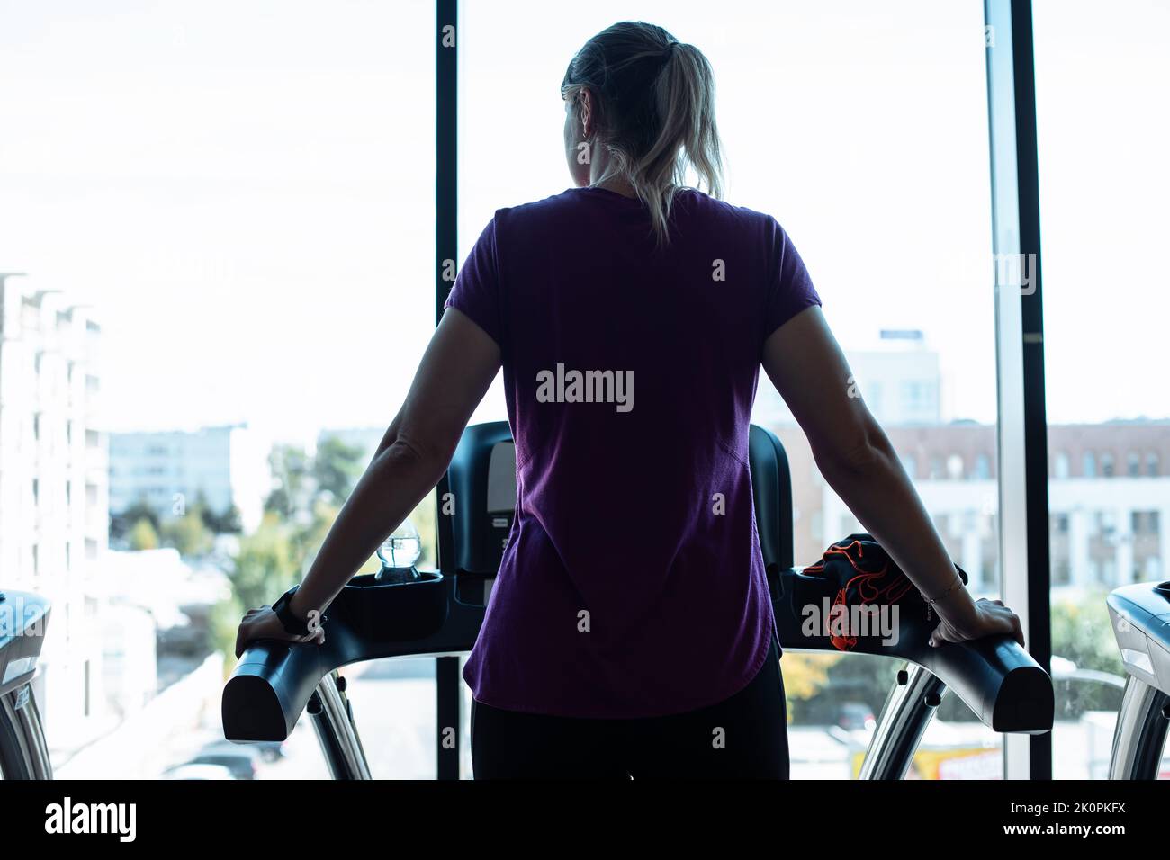 Plus size woman alone run on treadmill at fitness gym and look out window at city life, back view. Overweight girl do cardio workout on training Stock Photo