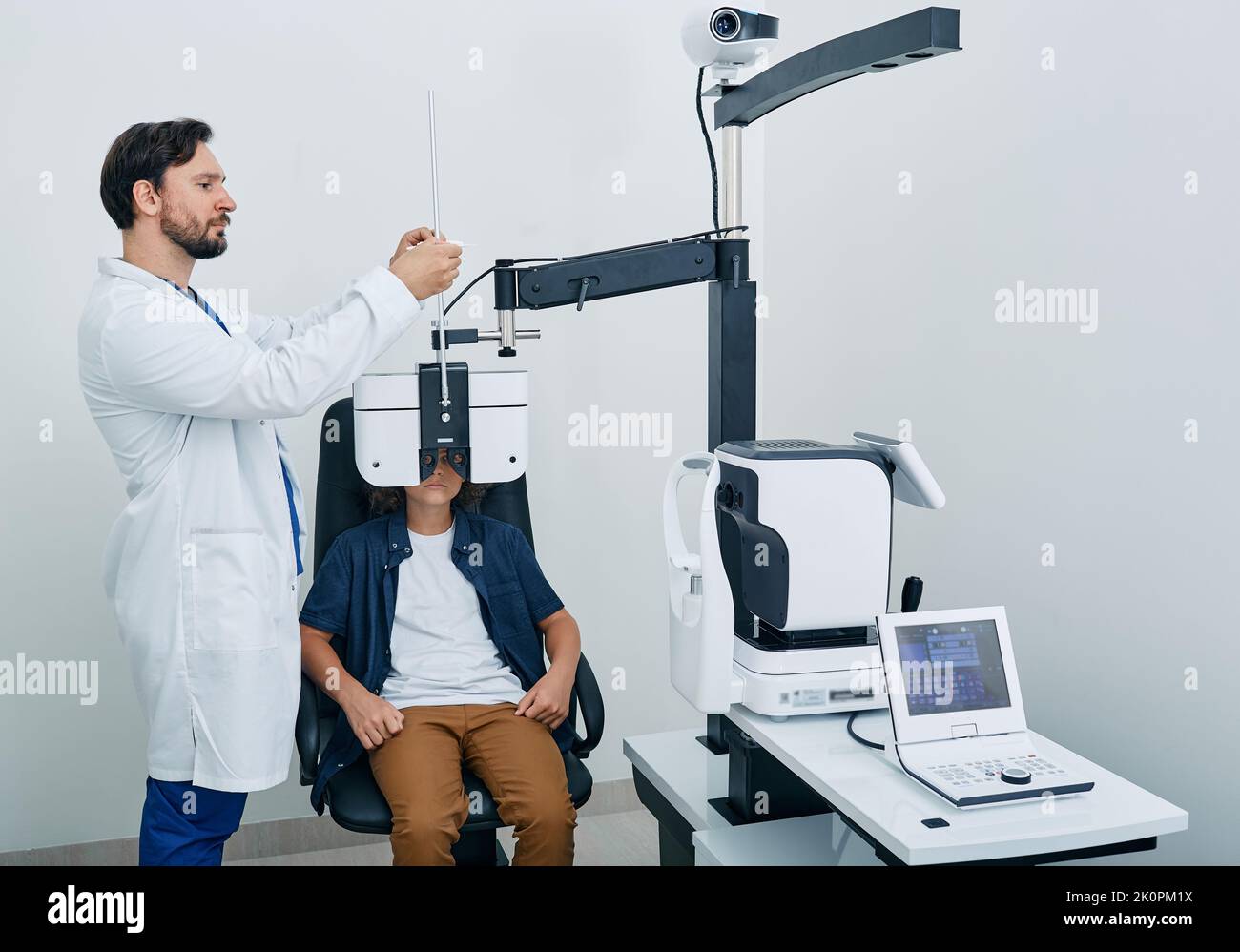 Optometrist doing vision test for curly boy patient using modern phoropter at ophthalmology clinic. Eye exam and vision diagnostic for children Stock Photo