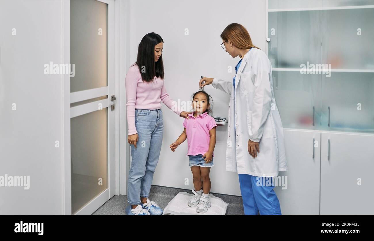 Pediatrician measures Asian female child's height during visit of child with her mother to medical clinic for consultation. Pediatrics Stock Photo