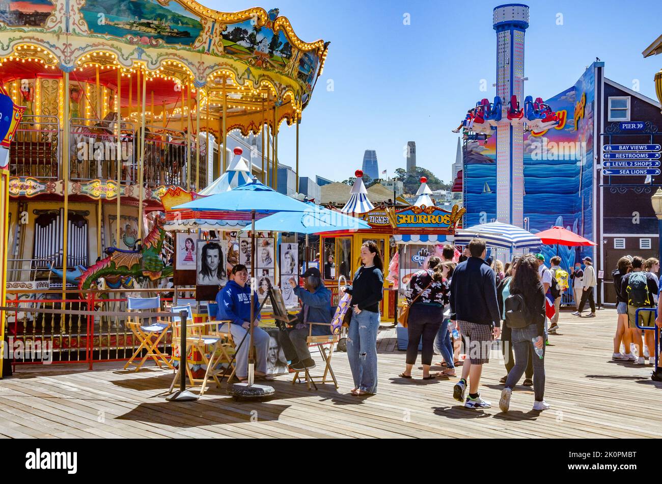 A view of people at Pier 39 with a carousel ride and an artist with a stall drawing caricatures on a summer's day with blue sky in San Francisco. Stock Photo