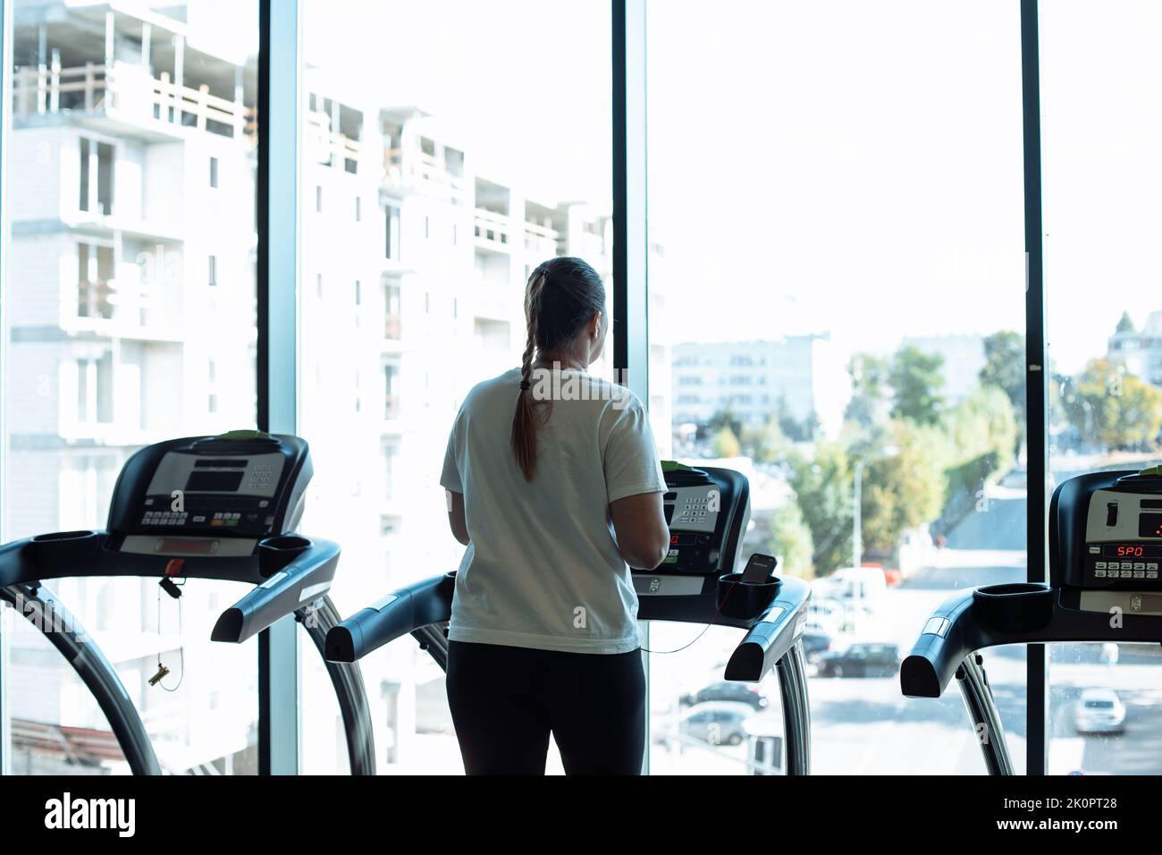 Fat woman run on treadmill at fitness gym and look out window at transport traffic and city life, back view. Overweight lady do cardio workout on Stock Photo