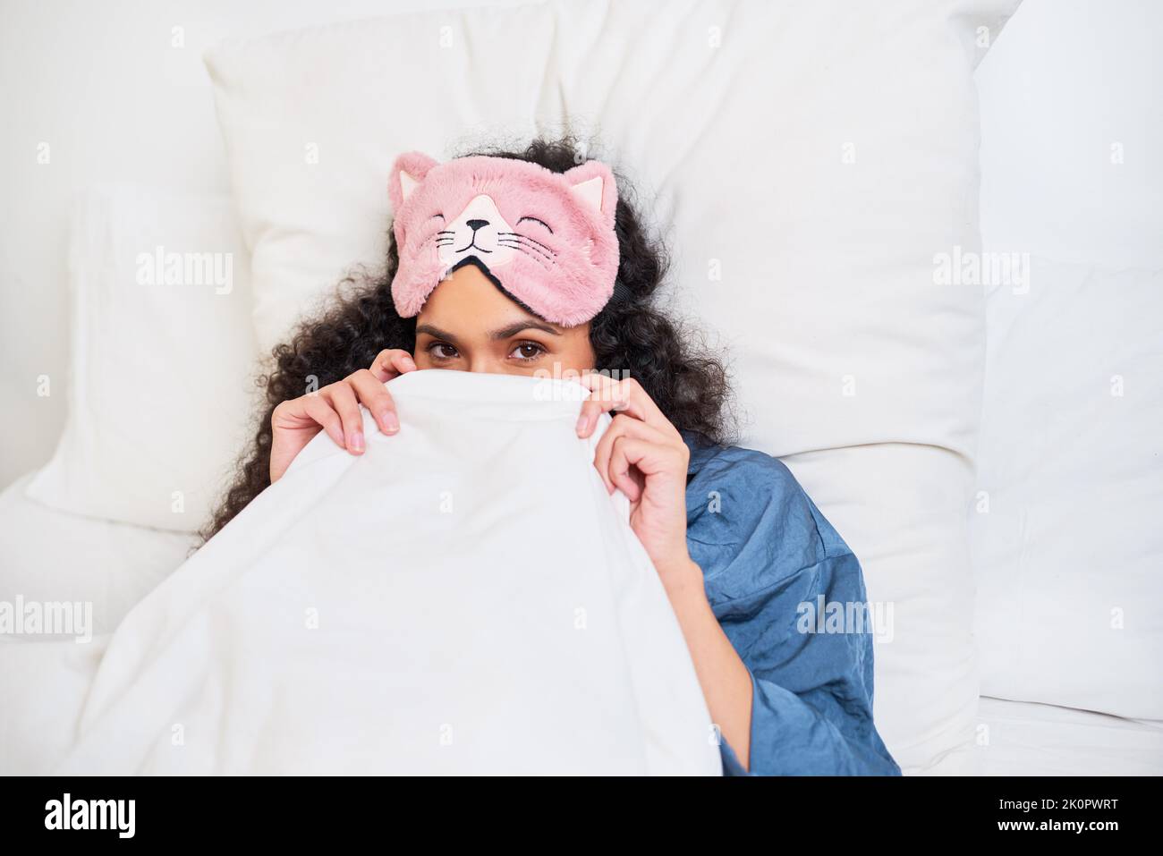 A young multi-ethnic woman hides under duvet in the morning with sleep mask on Stock Photo