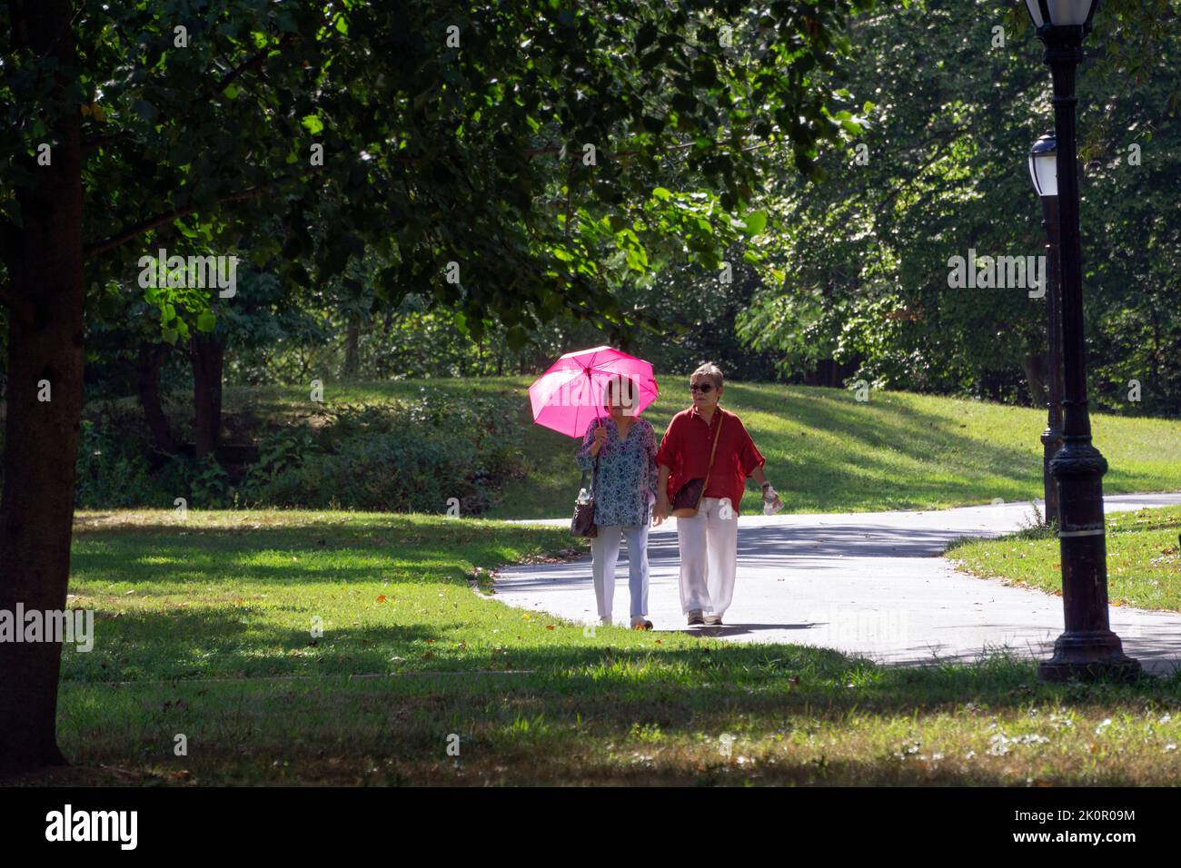 Two middle age Asian American women take an exercise walk on a sunny late summer day. One shields herself from the sun with a bright magenta umbrella. Stock Photo