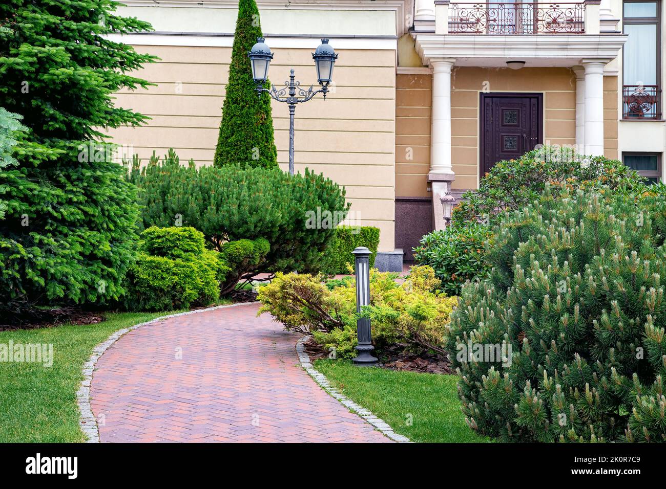 Backyard with bark mulched evergreens pine plants trees and bushes with needles and retro iron twin lantern and ground lighting near brick stone tile Stock Photo