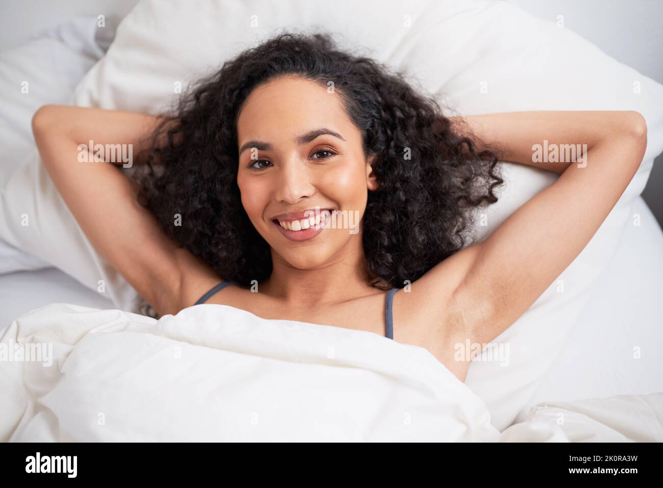 A young multiethnic woman lies with arms behind head in bed smiling