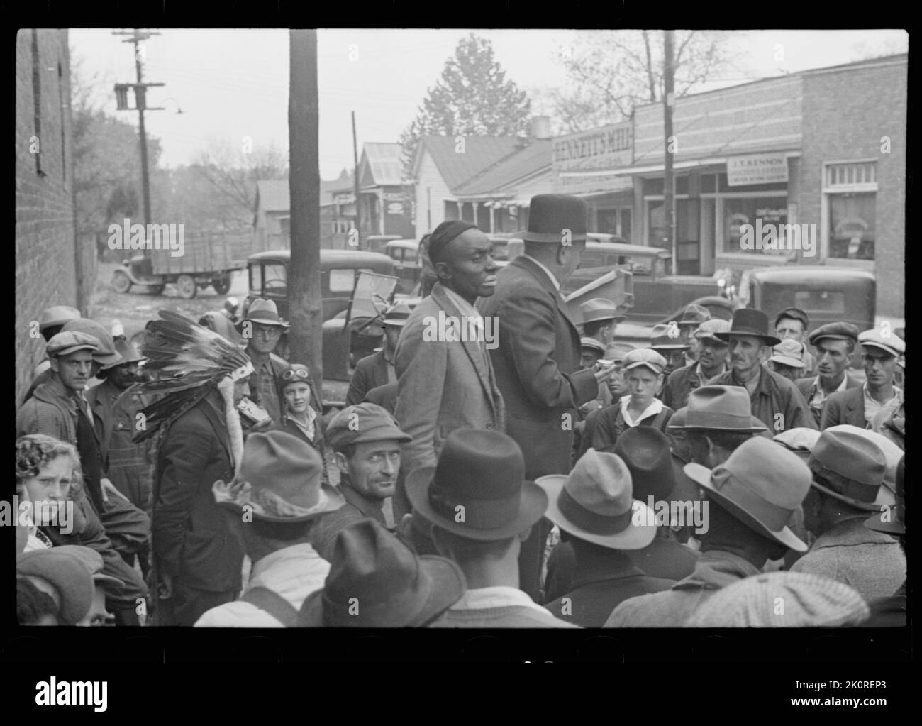 Medicine show draws a mostly white crowd listening to sales pitch delivered by a man (center, with face turned away) holding a tin. Other performers are an African-American in partial blackface (center) and a man wearing an Indian bonnet (lower left), , Huntingdon, TN, 10/1935. (Photo by Ben Shahn/FSA-OWI) Stock Photo