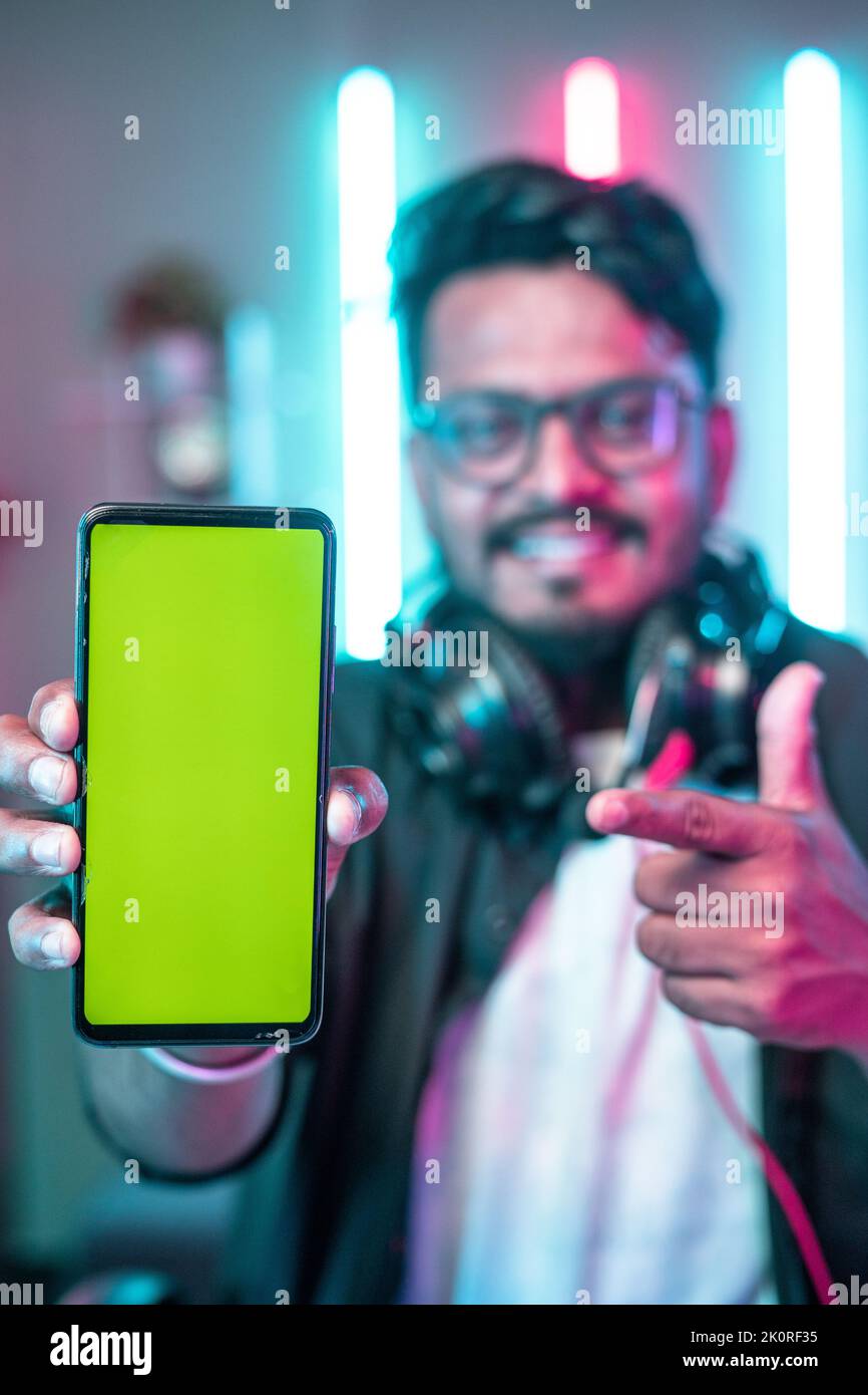 vertical shot of Professional gamer with headset showing green screen mobile phone by pointing finger while looking camera at home - concept of Stock Photo
