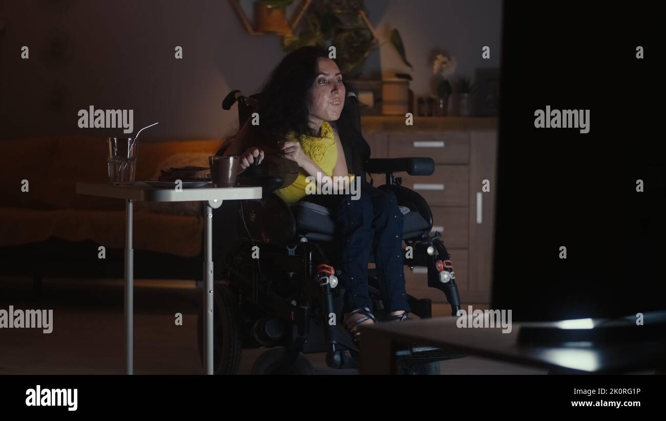 Woman with disability in a modern motorized wheelchair driving into living room and watching TV series in the evening while having dinner Stock Photo